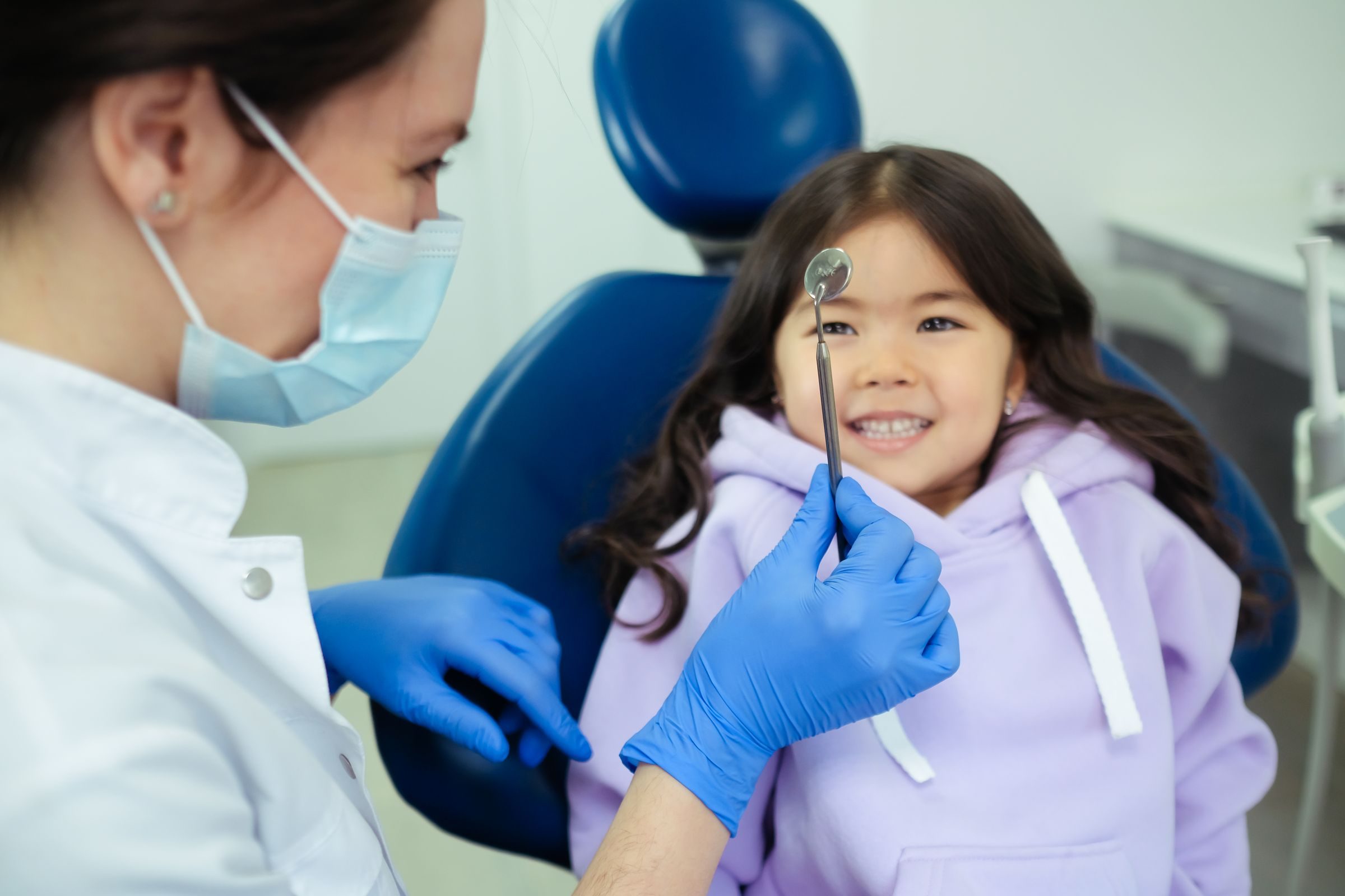 A young child smiling in a dental chair as the dentist gently shows them a tool — calm, friendly first dental visit