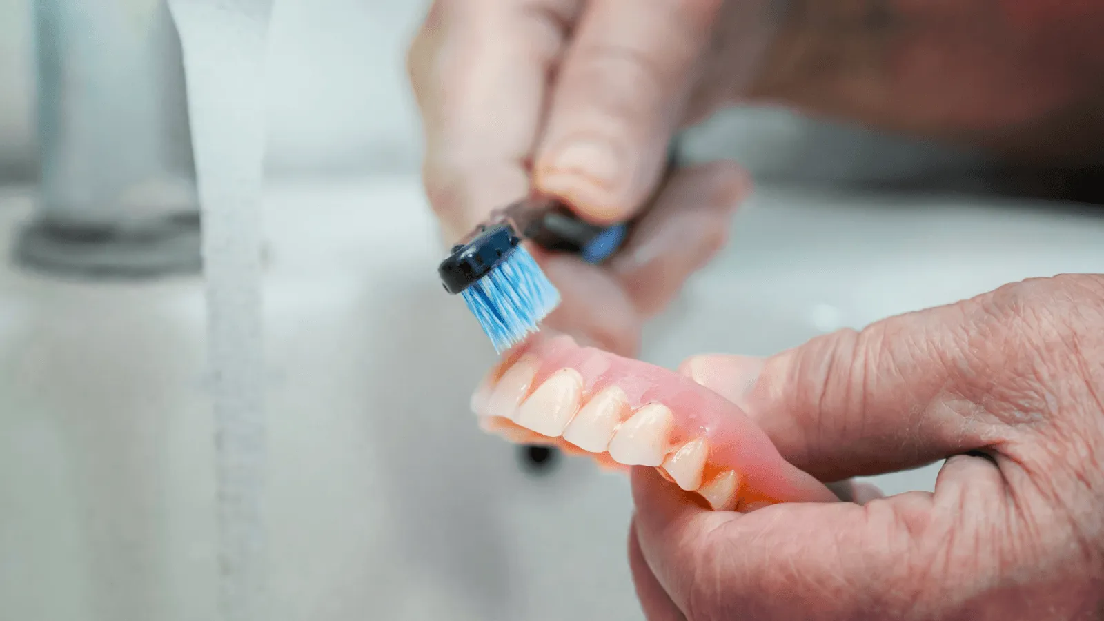Hands gently brushing a partial denture under a running tap, illustrating everyday denture care